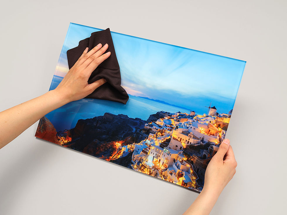 Hands cleaning a vibrant Santorini Acrylic Print with a cloth. The image is colorful, featuring brightly lit white buildings at dusk on a calm sea.