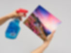 Hands holding a spray bottle with blue liquid and a colorful mountain landscape MetalPrint on a light gray background.
