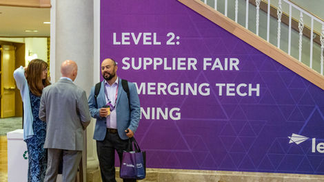 Conference attendees networking in venue corridor in front of a large a stairwell featuring "Level 2: Supplier Fair Emerging Tech" directional signage and triangular pattern design