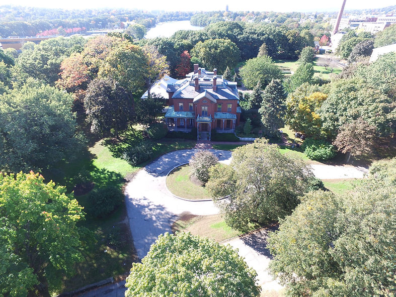 An aerial photo of the Commander's Mansion and grounds filled with many trees and the Charles River running along the back