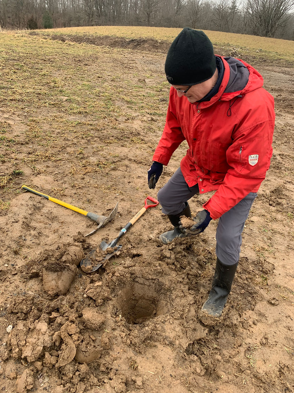 Man in red jacket digging holes in muddy field with shovel and spade. Overcast sky, barren trees in background. Boots and gloves worn.