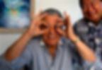 Salvador Assael smiling; holding dark pearls to his eyes, mimicking glasses. They're indoors with a colorful abstract painting on the wall.