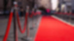 Red carpet lined with stanchions leads down a city street. Evening setting with blurred pedestrians and buildings in the background.