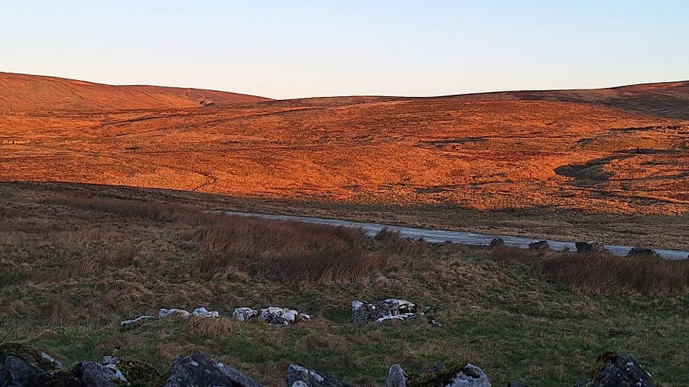 Stone wall in foreground, rolling hills bathed in orange sunset glow, under a clear blue sky. Serene and picturesque landscape.