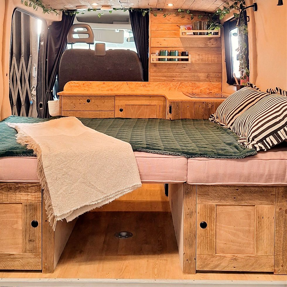 Cozy van interior with a wooden finish features a bed with green and striped cushions, beige throw, shelving with jars, and curtain accents.
