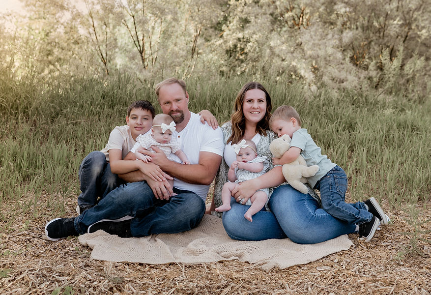 Family of six sits on a blanket in a grassy field; two baby girls, a toddler boy with a stuffed animal, another young boy, and parents smiling. Warm, sunny ambiance.
