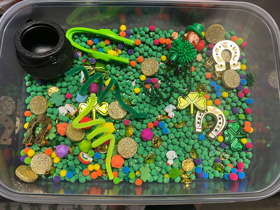 Plastic bin filled with colorful beads, gold coins, a black cauldron, green clovers, a toy leprechaun, and horseshoe charms. Festive mood.