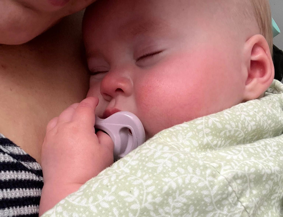 Baby sleeping peacefully with a pacifier, wrapped in a green patterned blanket, held against an adult's striped shirt. Calm mood.