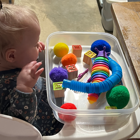 Colorful sensory toys and alphabet blocks in trays on a wooden table. Includes pom-poms, twisty tubes, and vibrant rings. Relaxed mood.