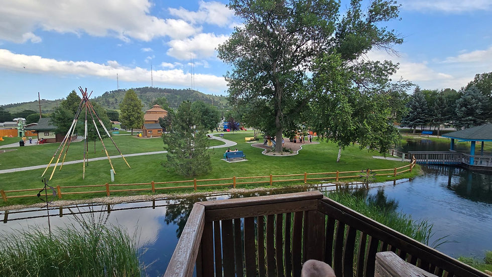 Park scene with teepee poles, a blue bench, and a tree under a partly cloudy sky. People relax on the grass near a pond and wooden deck.
