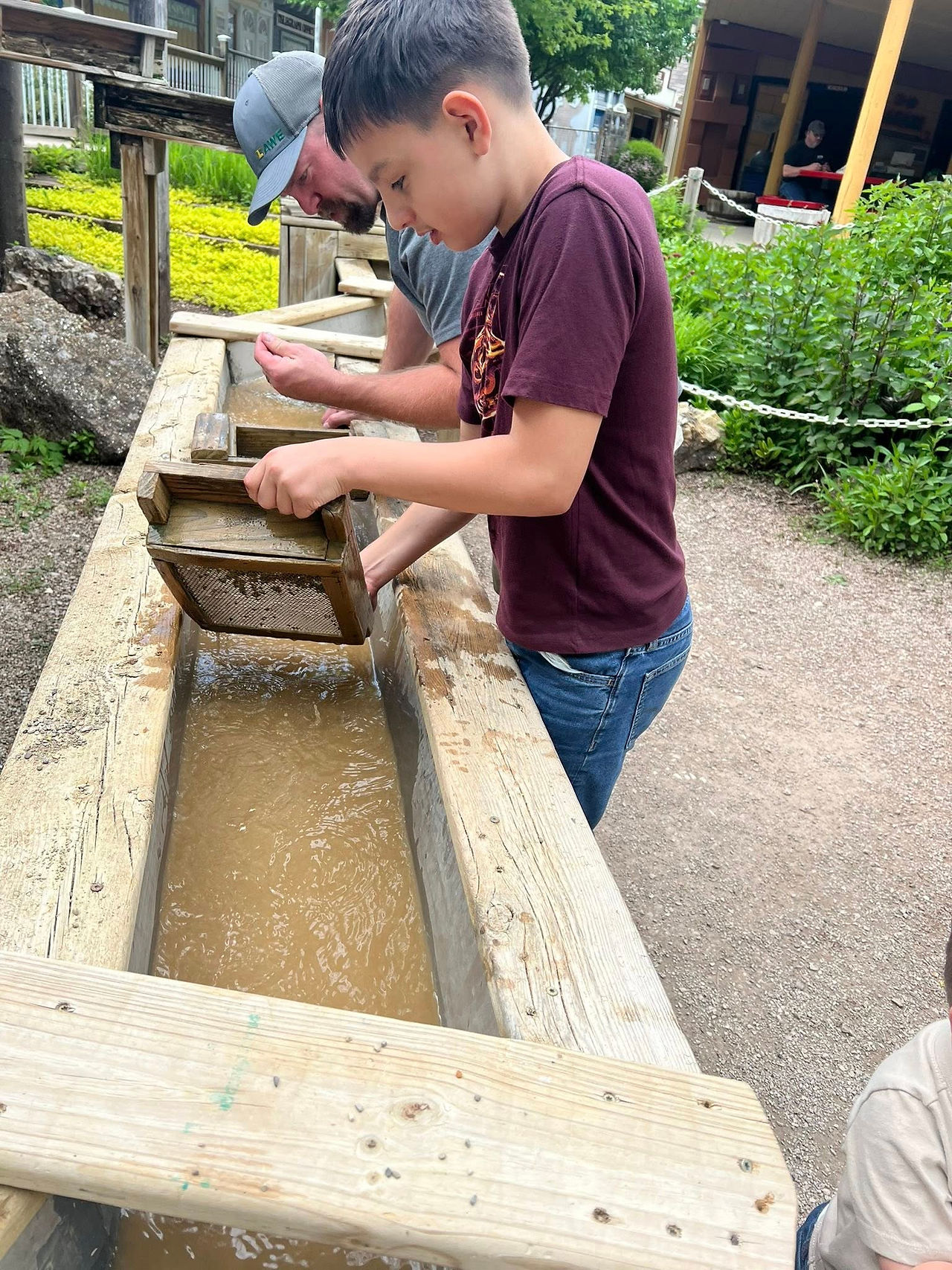Boy and man pan for gold in outdoor trough. Boy wears maroon shirt, man in grey cap. Greenery and wooden structures in the background.