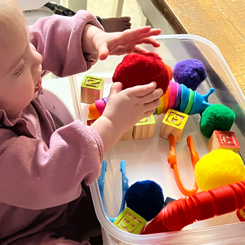 Colorful sensory toys and alphabet blocks in trays on a wooden table. Includes pom-poms, twisty tubes, and vibrant rings. Relaxed mood.