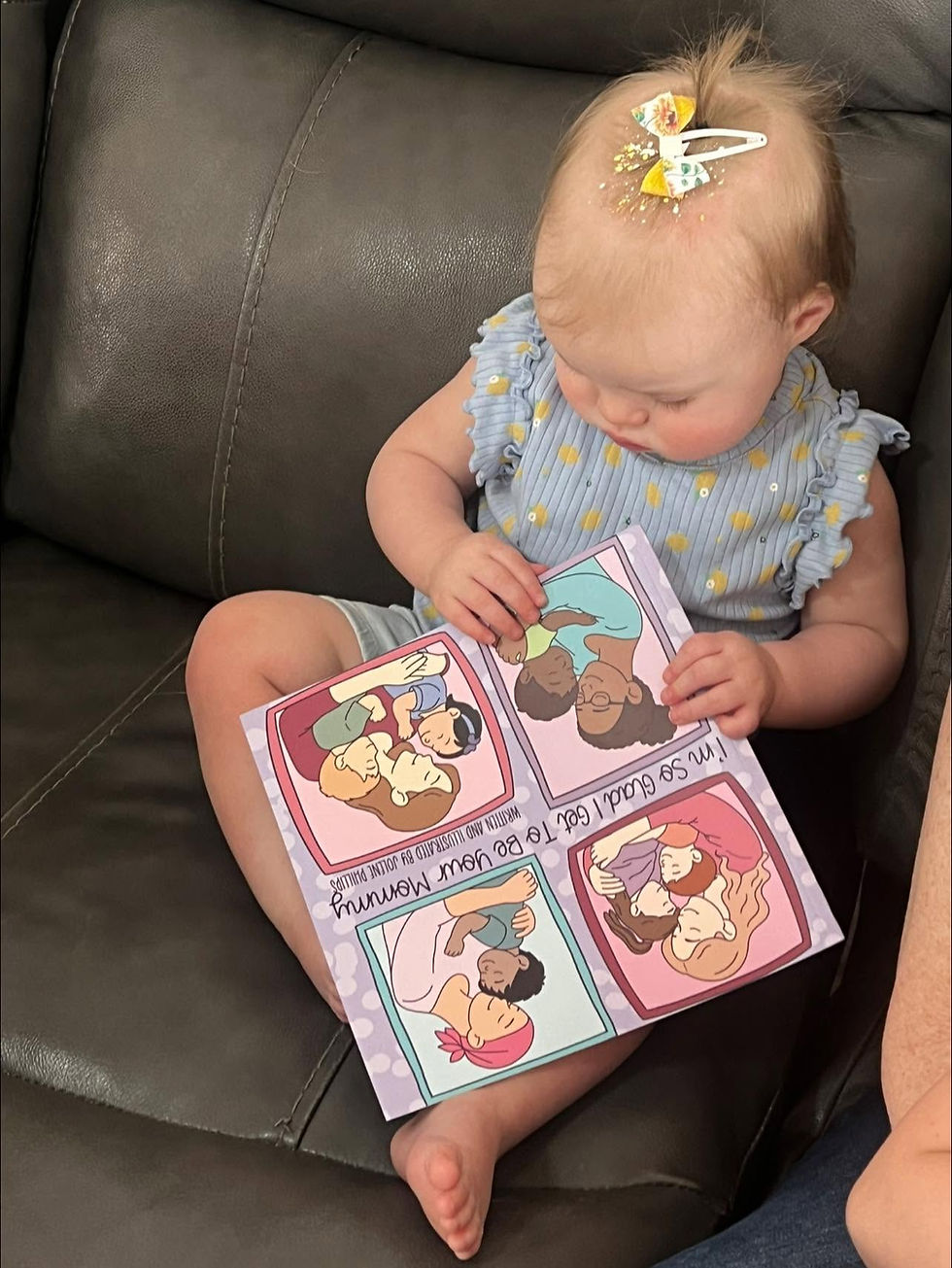 Baby girl with bow sits on a dark couch, looking at a colorful family book; text says I’m so glad I got to be your mommy.