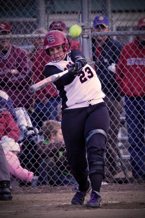 Softball player in a white and black uniform hits a ball during a game. A crowd in red watches from behind a chain-link fence.