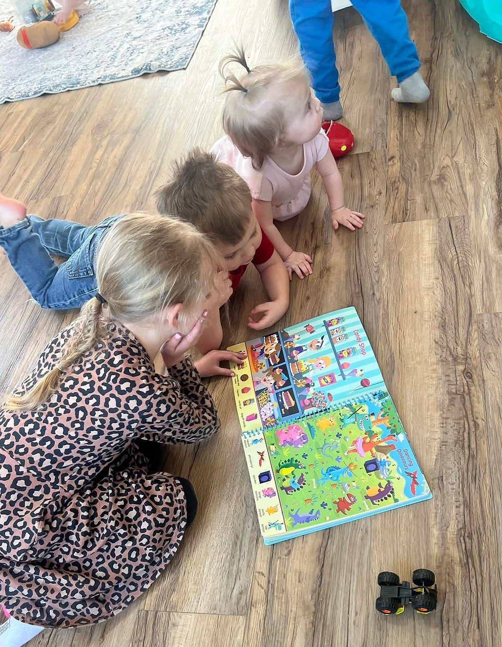 Toddlers on a wood floor gather around a colorful board book with candy shop and dinosaur pages, in a calm playroom.
