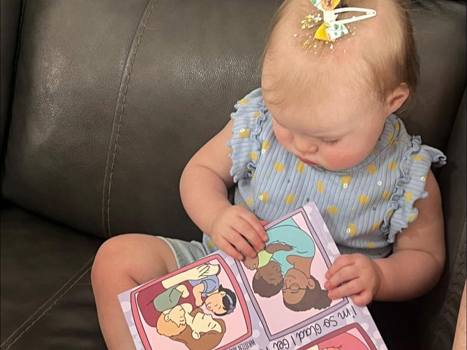Baby girl with bow sits on a dark couch, looking at a colorful family book; text says I’m so glad I got to be your mommy.