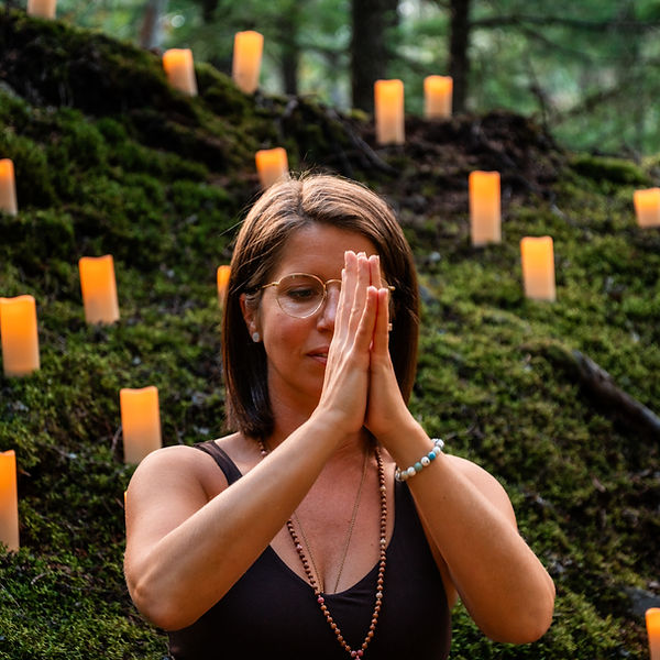 Katie from Peak Pose Yoga in Whistler seated on moss surrounded by candles, hands in Anjali Mudra (prayer gesture).