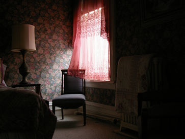 Light shines through a rose colored curtain onto a straight backed chair in a bedroom.