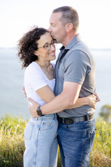 Seattle couple facing each other with arms around each other's waist smiling in front of the Pudget sound. Husband is kissing his wife's forehead.