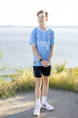 Teenage  boy in light blue shirt smiles at his family while standing in front of the Puget Sound at Discovery Park.