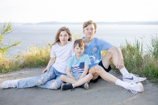 Three children, boy 7, girl 13, and boy 15, all are posing sitting down in the sand at Discovery Park with the Puget Sound behind them.