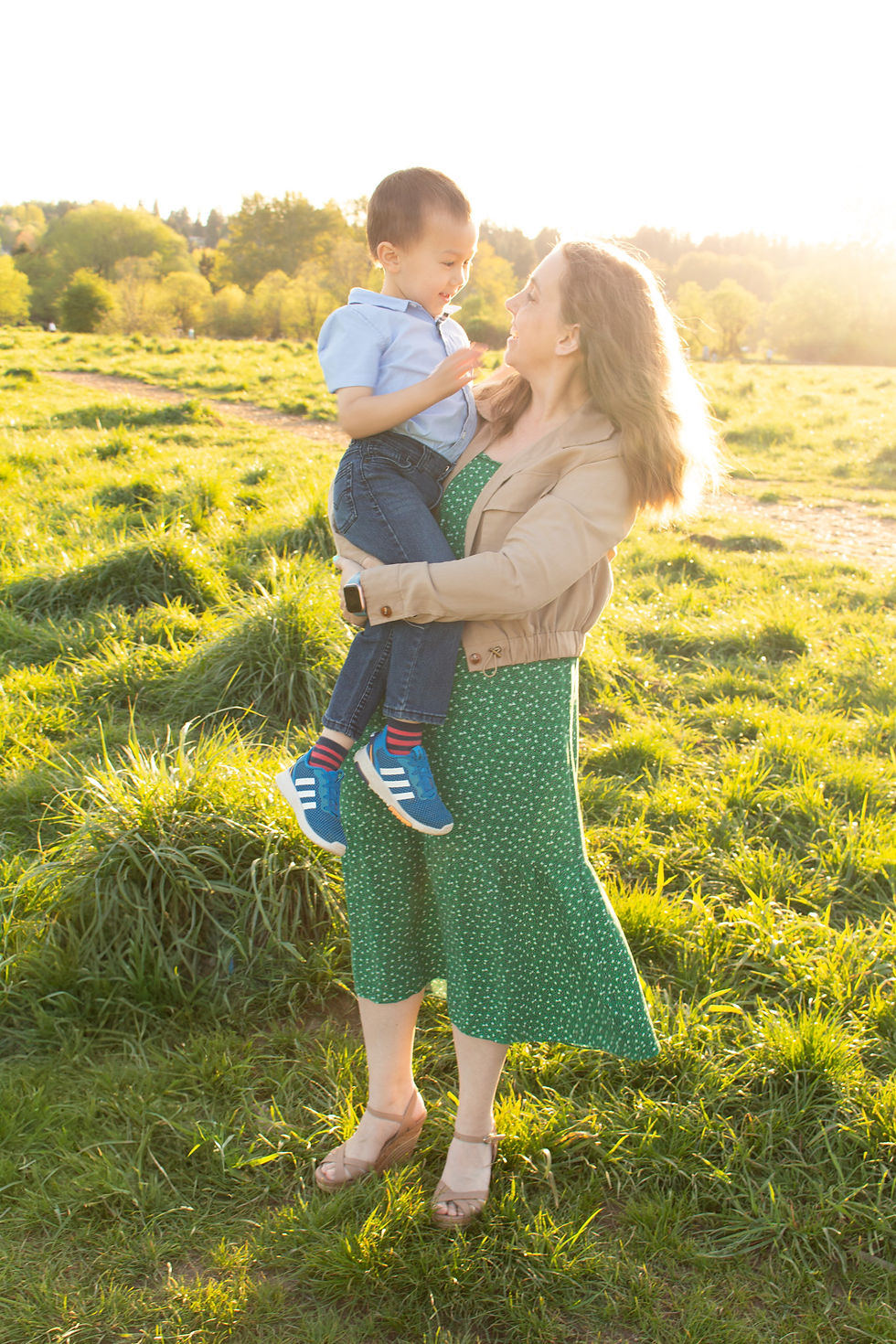 Mother in green dress holding son in light blue shirt and jeans in a sunny field for portraits