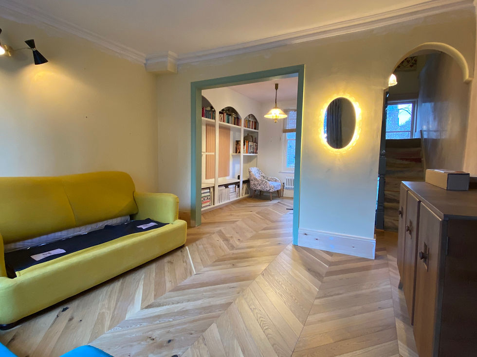 A cozy room with a yellow sofa, chevron wood flooring, and an archway leading to a lit room. An oval mirror glows on the wall.
