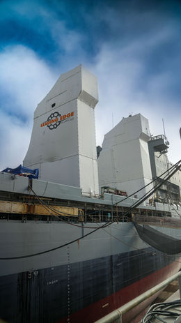 Leading Edge Scaffold’s white containment weather weapon a naval ship hull, with logo, cables, and safety netting under cloudy sky.