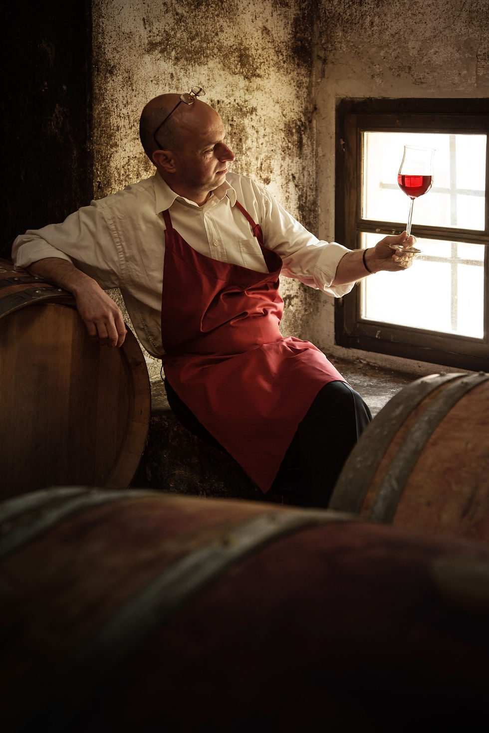 Man in a red apron examines a glass of red wine by a window in a rustic cellar with wooden barrels, creating a thoughtful mood.