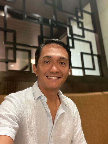 Smiling Ryan in a white shirt posing indoors. Geometric black patterns in the background. Warm lighting creates a cozy atmosphere.