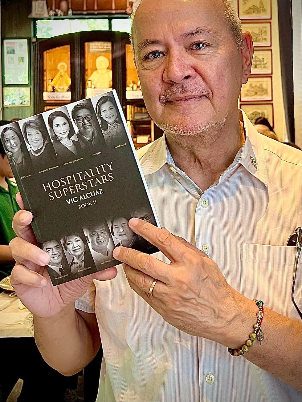 Vic Alquaz in a white shirt holding a book titled "Hospitality Superstars" with black-and-white portraits. Background has wooden decor and framed art.