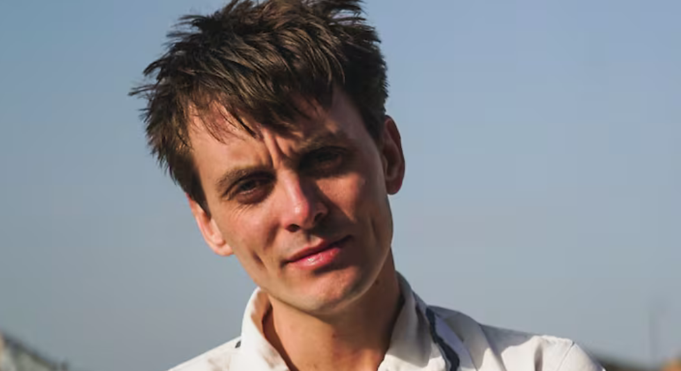 Man in white shirt outdoors, slightly smiling, with tousled hair against a clear blue sky. Bright lighting, relaxed mood.
