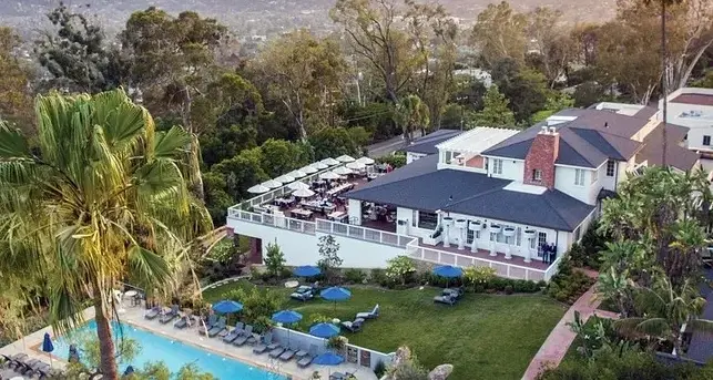 Aerial view of a resort with a pool, sun loungers, and blue umbrellas. A building with a patio and dining area overlooks lush trees and hills.