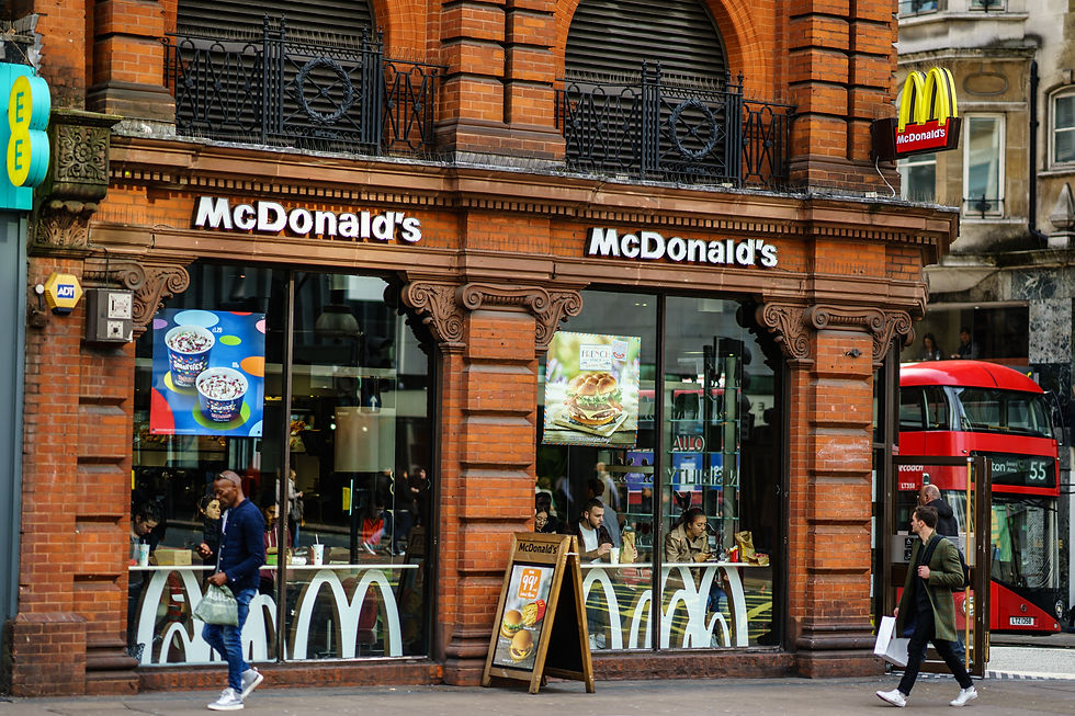 Urban McDonald's with red brick exterior. People pass by and dine inside. Visible signs, posters, and a red double-decker bus nearby.