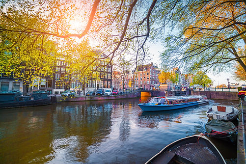 Canal scene in Amsterdam with a blue tour boat gliding by. Trees with green leaves hang over the water. Sunlight casts a warm glow.