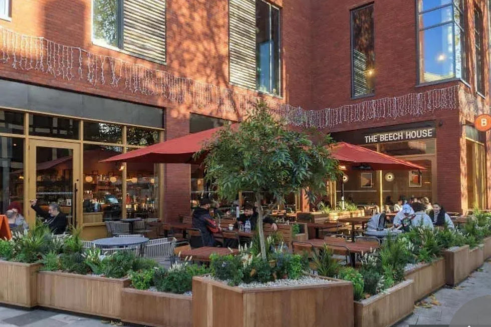 Outdoor seating at The Beech House with people dining under red umbrellas. Surrounded by greenery, set against a brick building. Cozy mood.