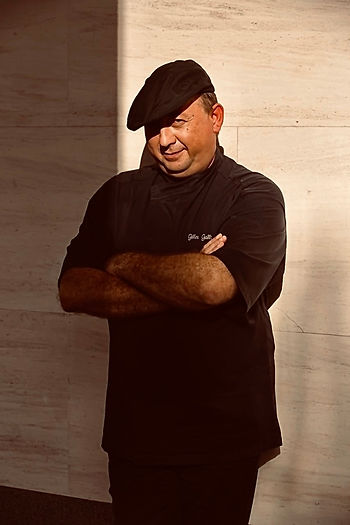 Gilles Galli in black outfit and beret, arms crossed, smiles in sunlight against a beige wall. Soft shadows enhance a relaxed mood.