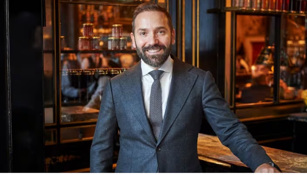 Smiling Baton Berisha in a gray suit stands in an elegant bar with glass shelves and warm lighting, creating a welcoming atmosphere.