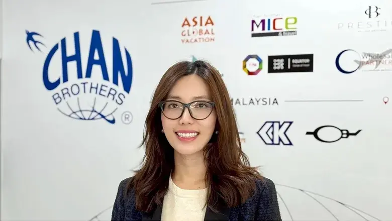 Smiling woman with glasses stands before a wall with travel brand logos, including Chan Brothers. Bright, professional setting.