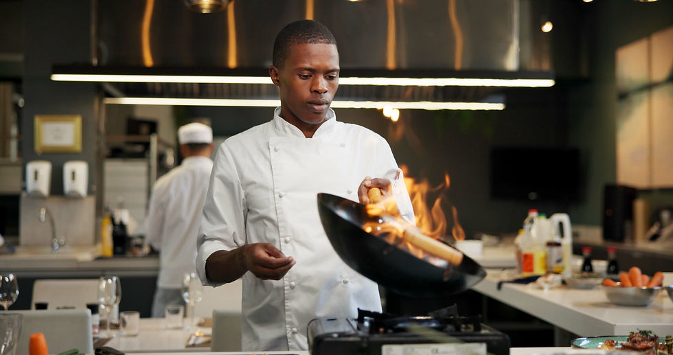 Chef in a white uniform tosses food in a flaming wok in a modern kitchen. Another chef works in the background. Stainless steel decor.