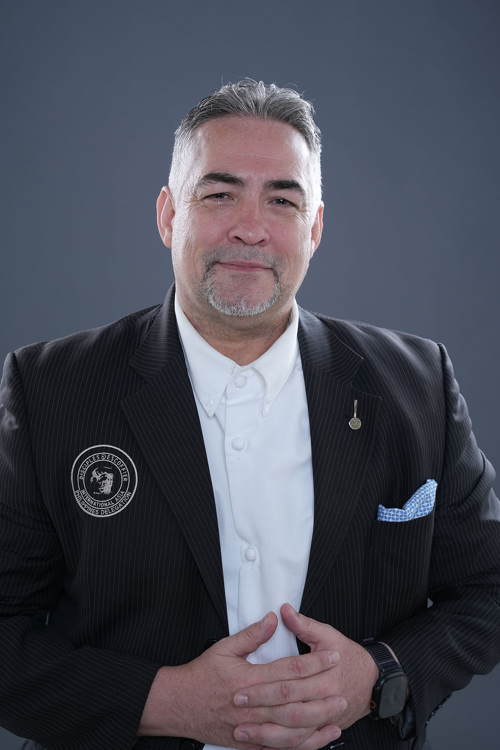 Gilles Galli in pinstripe suit with badge and pocket square, hands clasped, slight smile, against gray background. Formal and composed mood.