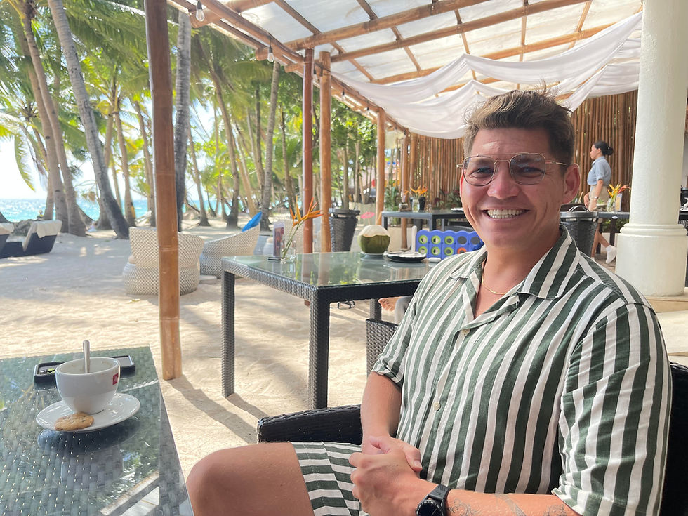Smiling person in a green striped shirt sits at an outdoor cafe with palm trees and sandy beach view. Coffee cup and biscuit on the table. Airy vibe.