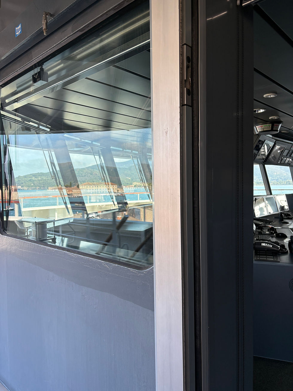 Ship bridge interior with dark control panels and windows overlooking a sunny bay and mountains
