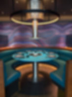 Futuristic dining area with a turquoise round booth table set for six, curved blue seats, and a vibrant wavy-patterned wall backdrop. 'Gunbae' Korean BBQonboard Virgin Voyages Brilliant Lady.