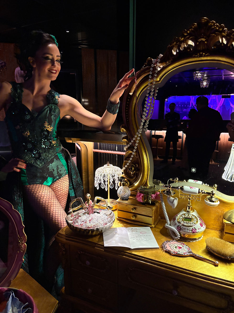 Woman in ornate costume poses joyfully by a vintage vanity with pearls, a pink phone, and makeup items. Dimly lit, elegant room.