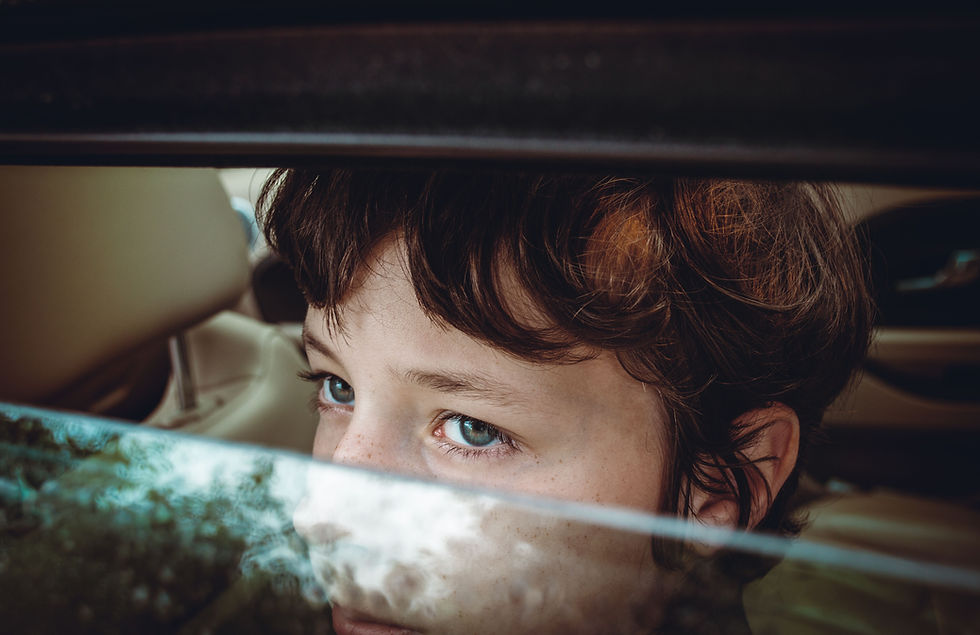 Child with blue eyes peeks through car window blinds, soft light and reflections, thoughtful mood.