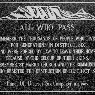 White text is placed on a black background within a white rectangle. The main block of text says: ‘All who pass: Remember the thousands of people who lived for generations in District Six and were forced by law to leave their homes because of the colour of their skins. Remember St. Mark’s Church and the community who resisted the destruction of District Six.’