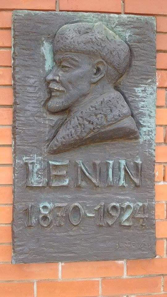 A textured bronze plaque situated against a backdrop of red bricks. The plaque shows a bas relief of the bust of a man with a small goatee in an ushanka style hat. Text below the man reads ‘Lenin 1870-1924’.