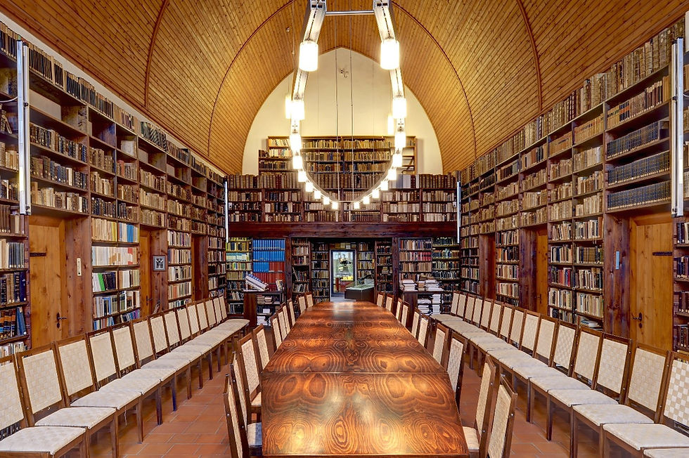 Long table in the centre of the shot with strip lights above. 13 chairs are on either side and the walls are covered in book cases. The ceiling meets in an arch at the top of the photo.