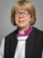 A smiling person wearing glasses and clerical attire with a purple and white collar, set against a plain grey background